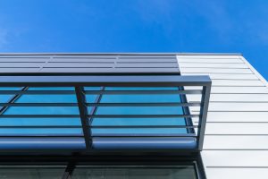 A modern building facade with horizontal metal slats and large glass panels against a clear blue sky. The structure features a minimalist design with a combination of glass and metal elements.