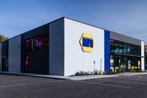 A modern building with a blue and gray facade featuring the NAPA Auto Parts logo. The front has large glass windows and doors with promotional signs. The parking lot is empty, and the sky is clear.