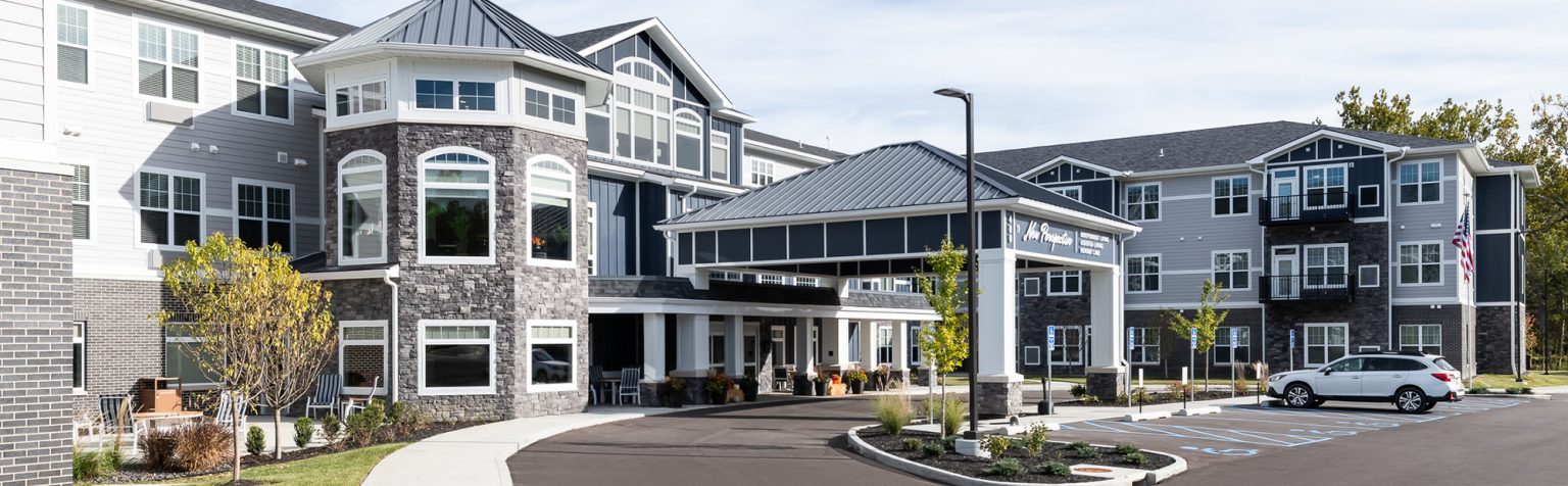 A large, two-story senior living facility with a modern facade, featuring gray stone accents and white siding. It has a covered entrance with a driveway, surrounded by landscaped greenery.