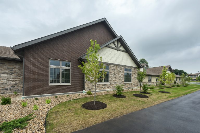 side view of building with brown siding, brick and cream colored siding, and landscaping with smaller trees