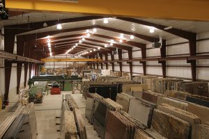 Interior of a large warehouse filled with slabs of stone or marble. The ceiling is high with exposed beams and overhead lights. The slabs are neatly stacked and categorized. A few machines and work areas are visible in the background.