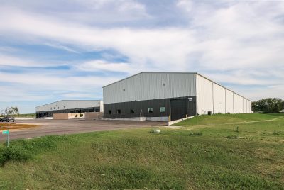 A large, modern warehouse building with a metal exterior. It has multiple loading docks and a spacious paved area and a grassy front lawn