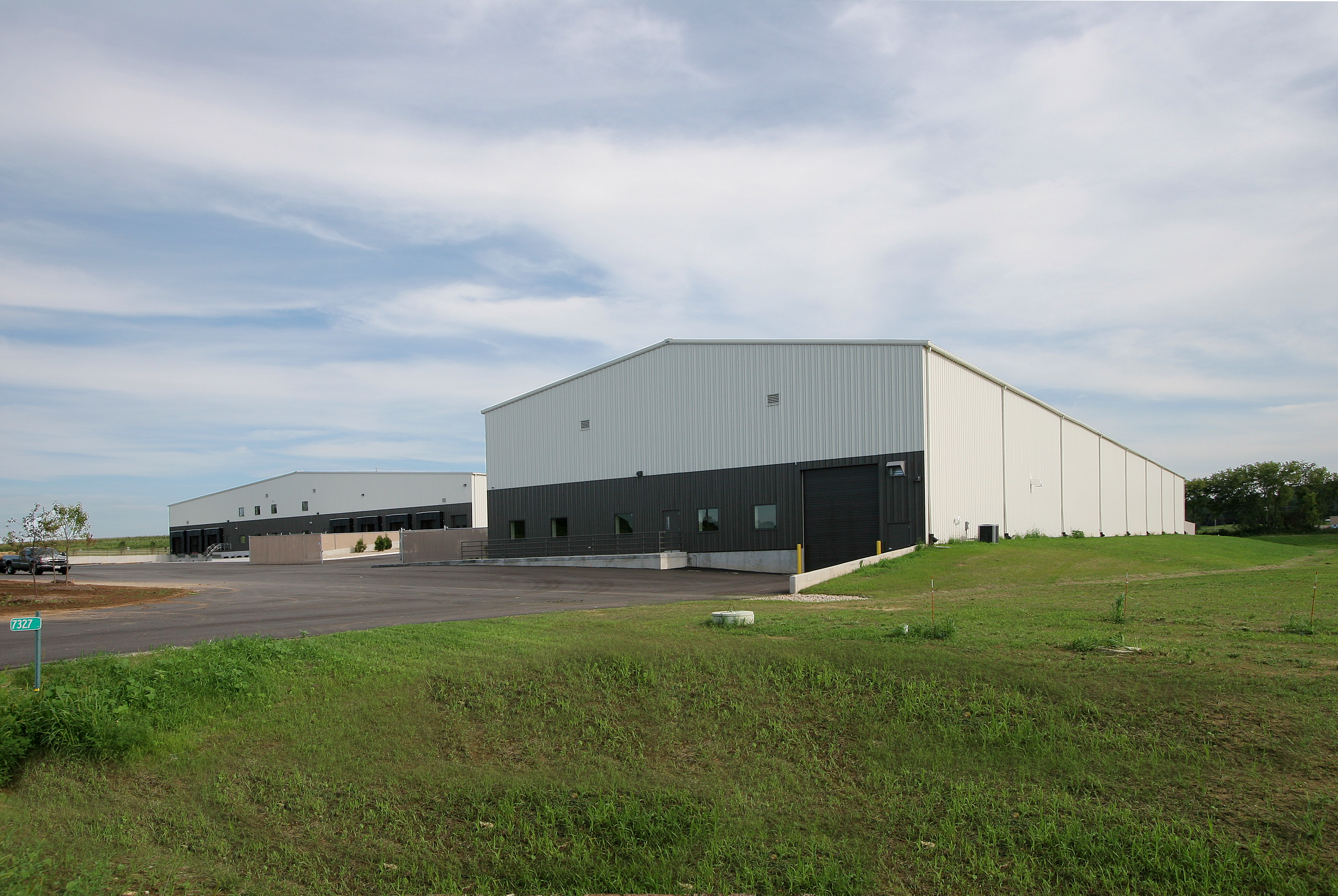 A large, white industrial warehouse with dark gray accents sits under a partly cloudy sky. It is surrounded by open grassy areas and a paved driveway leading to loading docks.