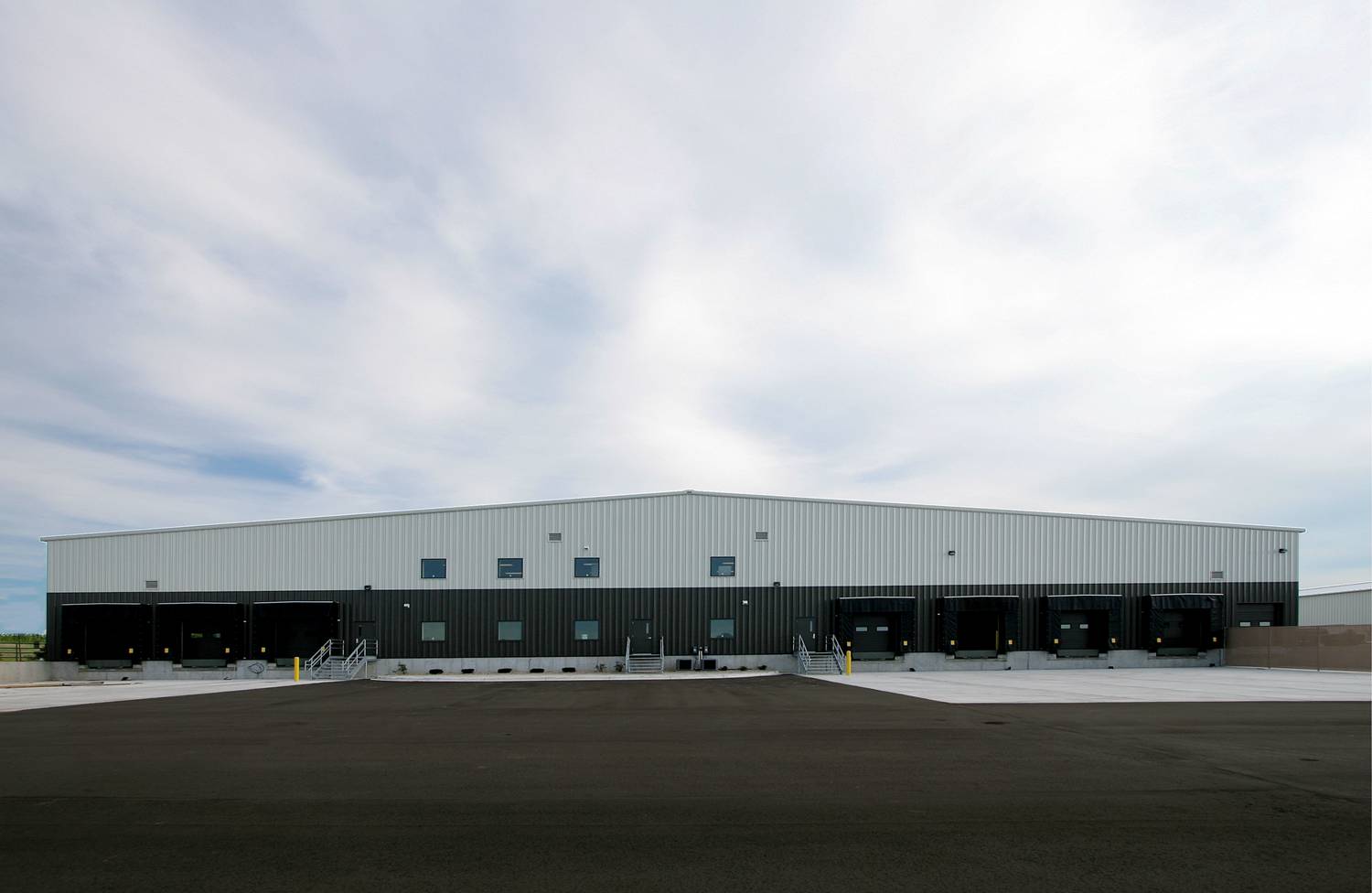 A wide-angle view of a large, modern warehouse building with a metal exterior. It has multiple loading docks and a spacious paved area in front. The sky is partly cloudy.