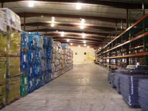 A spacious warehouse interior with tall stacks of assorted goods on pallets lining the left side and empty shelving units on the right. The concrete floor is clear, and overhead lighting illuminates the area.