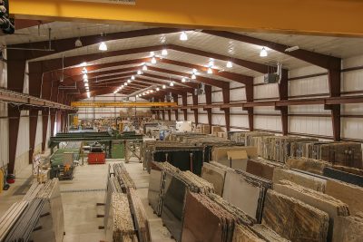 A large warehouse filled with neatly organized slabs of stone and granite. Overhead lights illuminate the spacious interior with rows of variously colored stone slabs stacked vertically. Industrial equipment is visible in the background.