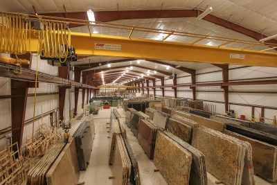 A large Industrial warehouse filled with neatly organized slabs of stone and granite. Overhead lights illuminate the spacious interior with rows of variously colored stone slabs stacked vertically.