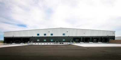 A wide-angle view of a large, modern warehouse building with a metal exterior. It has multiple loading docks and a spacious paved area in front. The sky is partly cloudy.