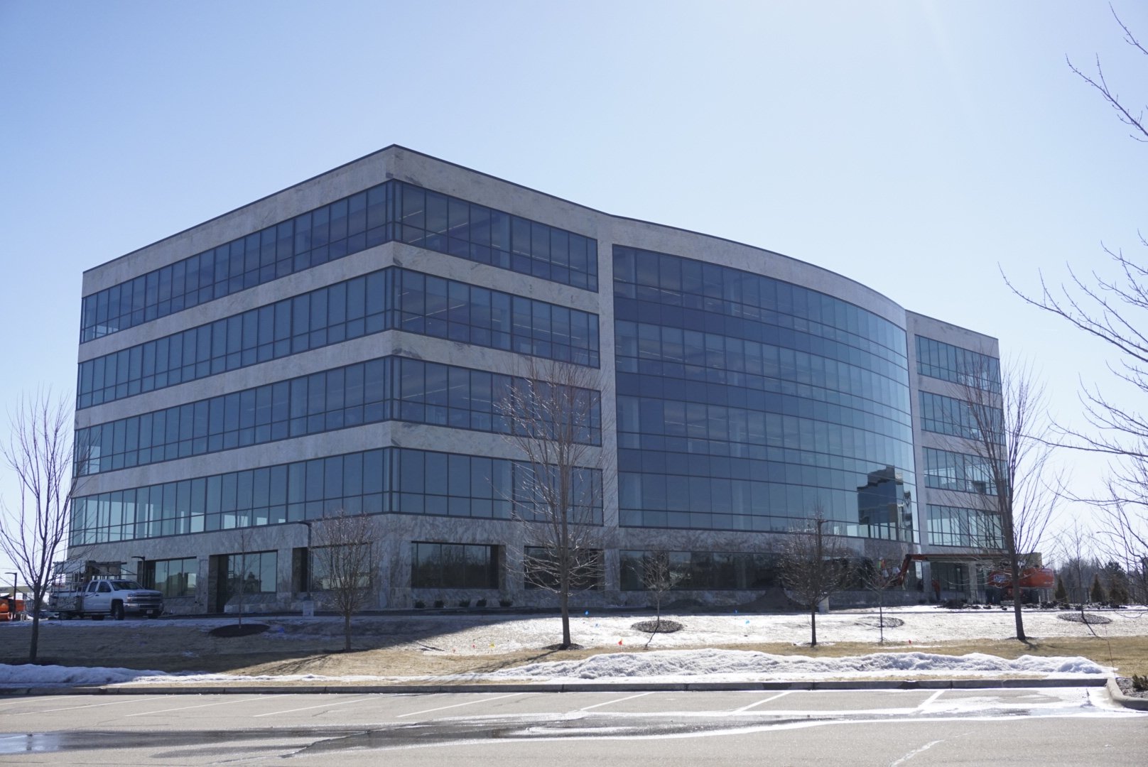 A large, modern office building with a glass facade and four stories, situated on a slightly snowy landscape. The sky is clear, and the building is surrounded by bare trees and a partially snow-covered lawn. There are a few parked vehicles nearby.