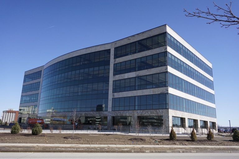 glass and stone office building on a clear day. The building has large, reflective glass windows and a sleek facade. The surrounding area includes a construction site and small landscaped trees.