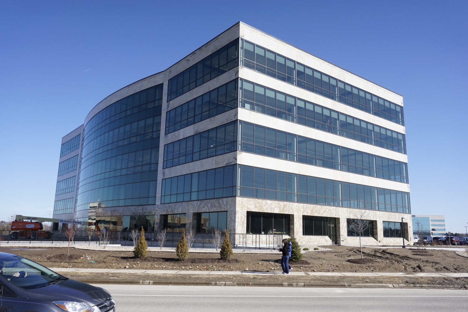 A large, modern office building with a curved design and extensive glass windows under a clear blue sky. The building is situated along a road with a few cars and people passing by. The surrounding area is under minor landscaping development.