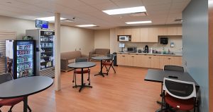 A break room with light wood flooring and neutral walls features a seating area with tables and chairs, a kitchenette with a microwave, coffee maker, and sink. A vending machine filled with snacks and beverages is near a small sofa. Ceiling lights illuminate the space.