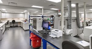 A modern laboratory with various scientific equipment and computers on workstations. The room has white cabinets, a black chair, and a red waste bin. The lab is well-lit with overhead fluorescent lights and has a clean and organized appearance.