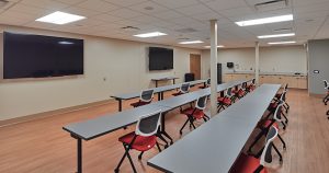 A modern classroom with long gray tables arranged in rows, red and black chairs, and multiple large flat-screen monitors on the beige walls. The wooden floor adds a warm touch, and the room is brightly lit by ceiling lights. The back wall has cabinets and a sink.