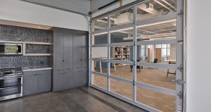 A modern office interior features a kitchen area with dark cabinets and a tile backsplash on the left. The room is divided by a large glass garage door. On the right, there's an open workspace with bookshelves and a whiteboard in the background.