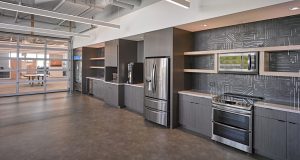 A modern office kitchen with stainless steel appliances including a fridge, microwave, stove, and dishwasher. The kitchen features dark wood cabinets, an abstract patterned backsplash, open shelves, and a polished concrete floor. A glass-walled meeting room is visible in the background.