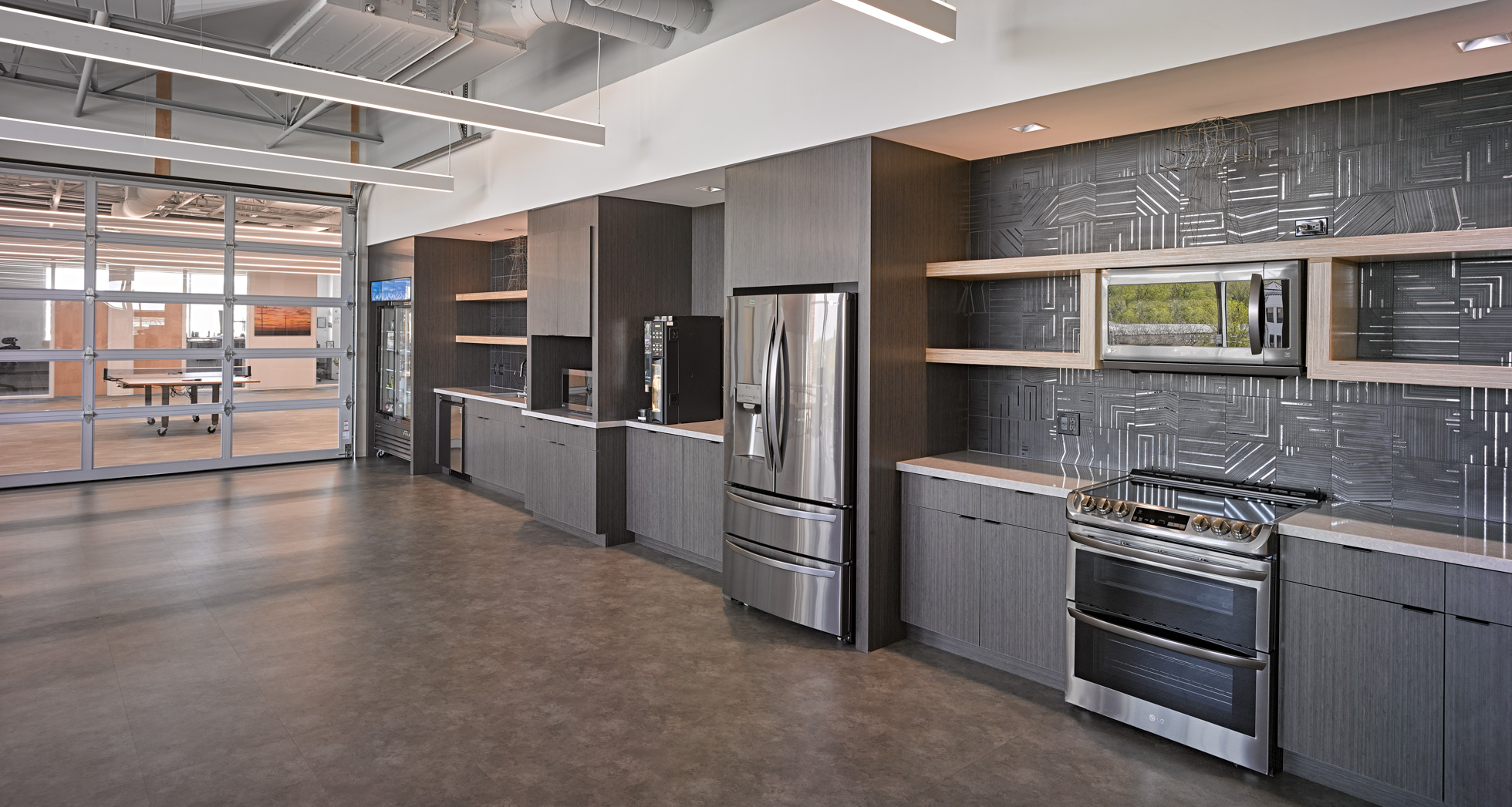 A modern office kitchen with stainless steel appliances including a fridge, microwave, stove, and dishwasher. The kitchen features dark wood cabinets, an abstract patterned backsplash, open shelves, and a polished concrete floor. A glass-walled meeting room is visible in the background.