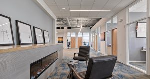 Modern office space featuring a cozy seating area with two black leather chairs facing a sleek, wall-mounted electric fireplace. The hallway is lined with framed artwork and leads to various glass-walled office rooms. Bright overhead lighting illuminates the area.