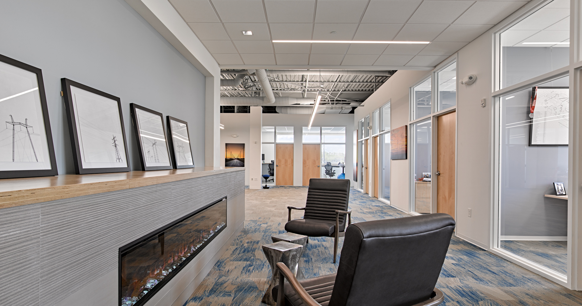 Modern office space featuring a cozy seating area with two black leather chairs facing a sleek, wall-mounted electric fireplace. The hallway is lined with framed artwork and leads to various glass-walled office rooms. Bright overhead lighting illuminates the area.