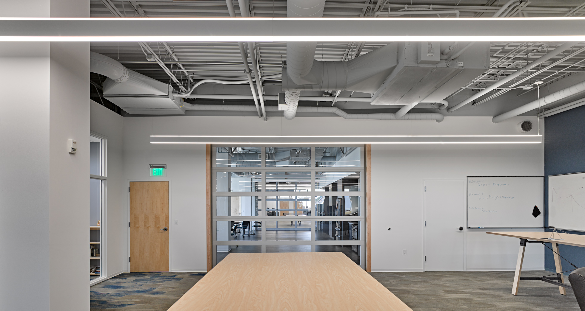 A modern office interior with an exposed ceiling, light gray walls, and a wooden door on the left. The room has long rectangular tables, a large window with blinds, a whiteboard on the right wall, and a glass partition leading to another office space in the background.