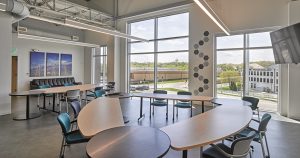 Modern common area with large windows and natural light, featuring curved tables arranged in a semicircle, black and teal chairs, a sectional sofa, and a flat-screen TV mounted on the wall. Wall art and decorative hexagonal tiles enhance the bright, open space.