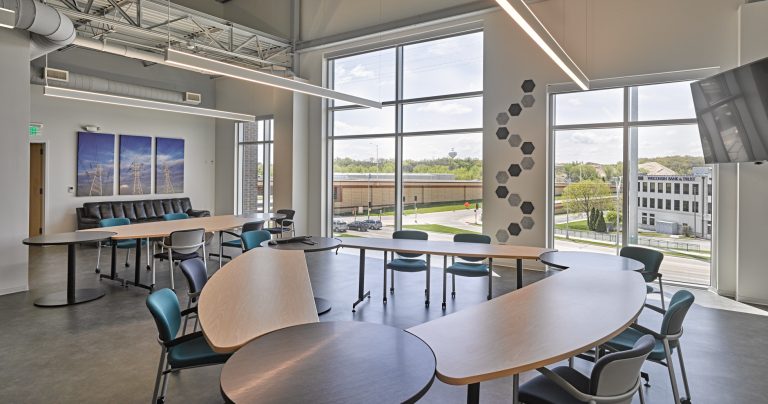 Modern common area with large windows and natural light, featuring curved tables arranged in a semicircle, black and teal chairs, a sectional sofa, and a flat-screen TV mounted on the wall. Wall art and decorative hexagonal tiles enhance the bright, open space.