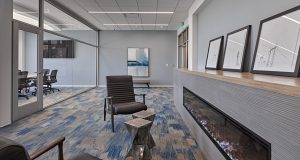 A modern office lounge with blue-patterned carpeting, black leather chairs, a small wooden side table, and wall art. A linear fireplace with a wooden mantel sits on one side, and in the background, there is a glass-walled conference room with several chairs.