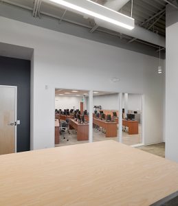 A modern office with wooden desks, computers, and ergonomic chairs. The room is viewed through large glass windows from an adjacent open area. Fluorescent lights illuminate the space, and the ceiling displays visible infrastructure and ducts.