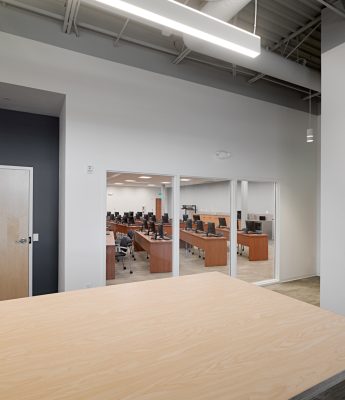 A modern office with wooden desks, computers, and ergonomic chairs. The room is viewed through large glass windows from an adjacent open area. Fluorescent lights illuminate the space, and the ceiling displays visible infrastructure and ducts.