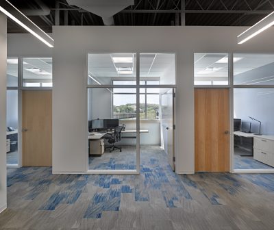 A modern office space with three enclosed glass-walled offices. Each office is equipped with desks, chairs, and monitors. The area outside the offices has blue and gray carpet. Large windows in the offices allow natural light to illuminate the interiors.