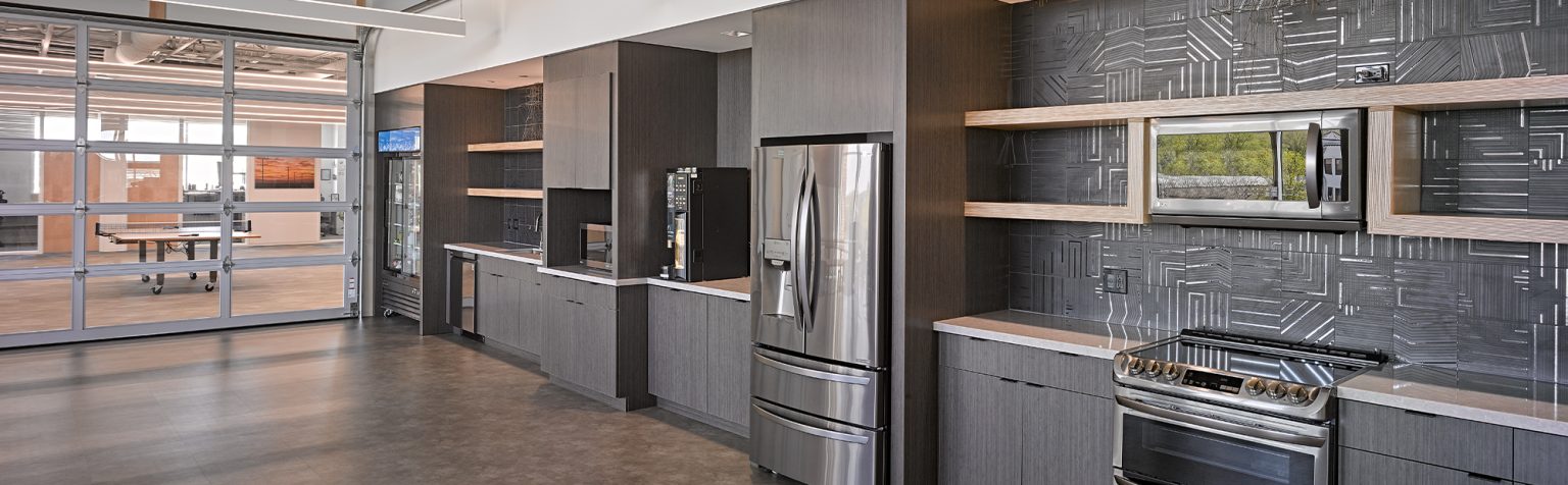 office kitchen with stainless steel appliances including a fridge, microwave, stove, and dishwasher. The kitchen features dark wood cabinets, an abstract patterned backsplash, open shelves, and a polished concrete floor. A glass-walled meeting room is visible in the background.