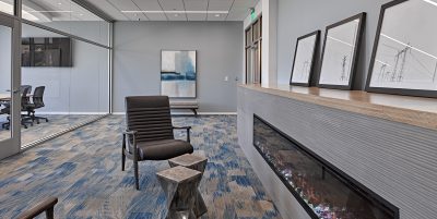 office lounge with blue-patterned carpeting, black leather chairs, a small wooden side table, and wall art. A linear fireplace with a wooden mantel sits on one side, and in the background, there is a glass-walled conference room with several chairs.