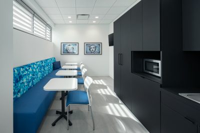 A modern office lunchroom with blue cushioned bench seating and white chairs around small white tables. The room has black cabinets, including a microwave, a sink, and two framed artworks on the wall. A window with blinds lets in natural light.