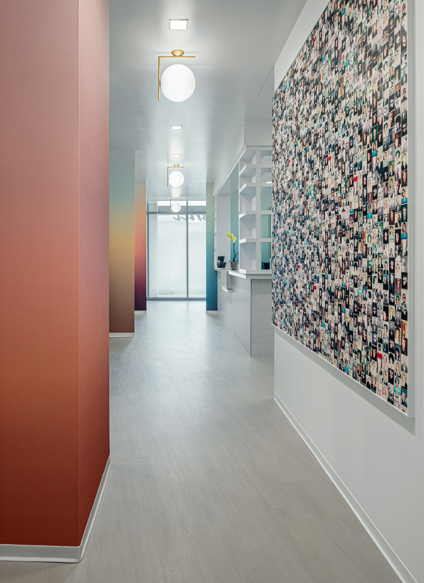 A modern hallway with light wood flooring and a gradient from red to beige on the left wall. A large collage made up of numerous small photos covers the entire right wall. At the hallway's end is a minimalist reception desk with shelves and a small potted plant.