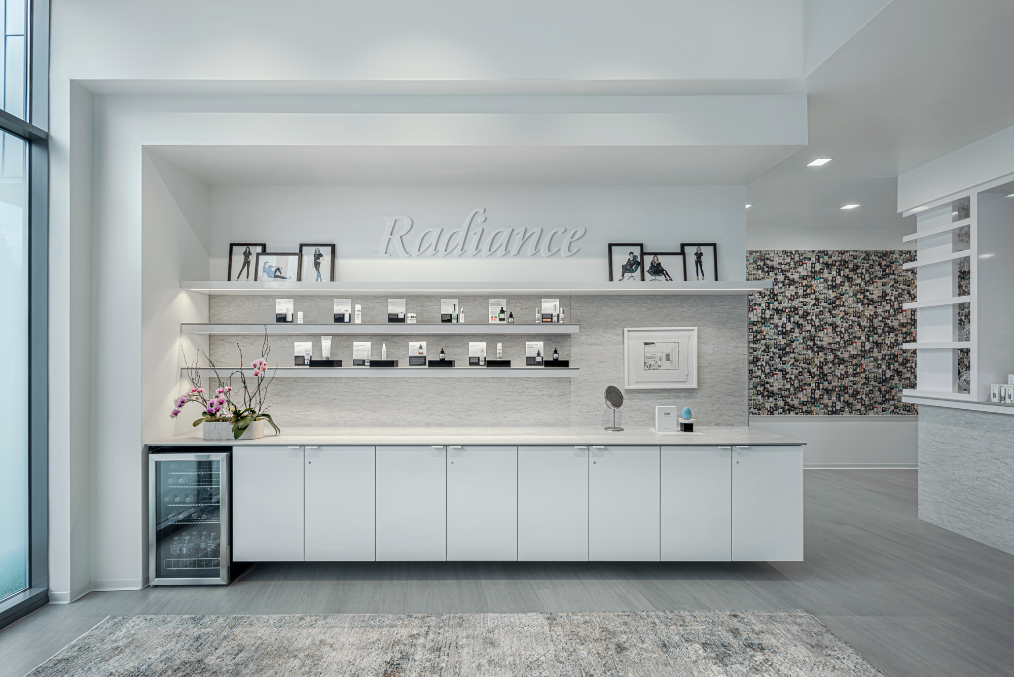 A modern, minimalist skincare display area with white cabinetry and countertops. Shelves above showcase skincare products and framed pictures. "Radiance" is written in cursive on the wall. A small fridge is on the left, and a floral-patterned wall is visible in the background.