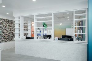 Modern reception area with white shelves displaying various skincare products. The background features a mosaic art wall and a multicolored gradient wall. Two computer workstations are visible behind the counter, with a small plant adding a touch of greenery.