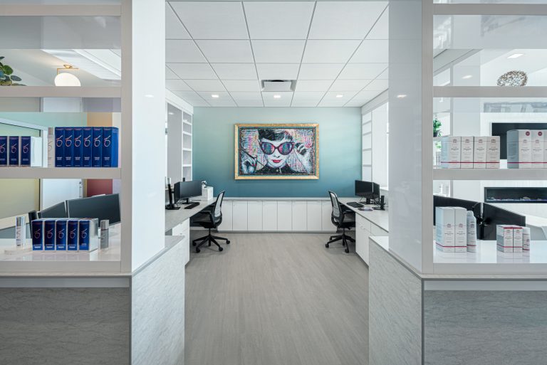 A modern office space with white shelves displaying various skincare products. Two workstations with computers face inward, and a large, colorful portrait of a woman with sunglasses is hung on the blue accent wall in the background.