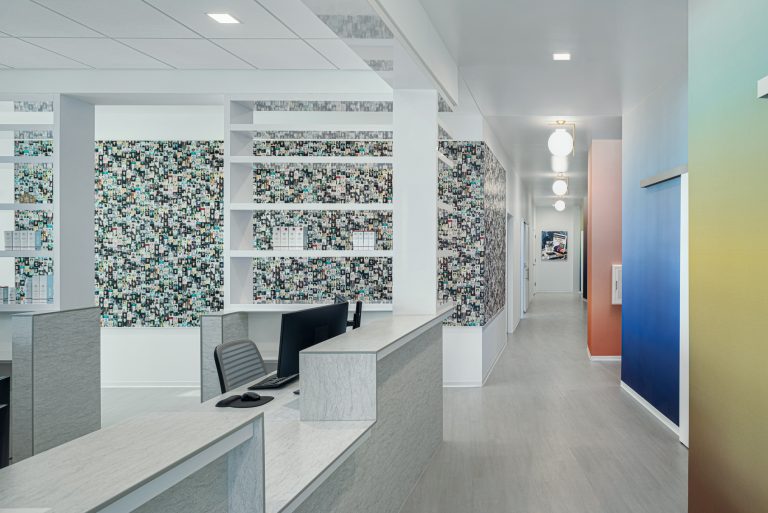 A modern, brightly lit office hallway with colorful mosaic walls and open shelving. The space includes a reception desk with a computer and chair. Hanging spherical lights line the hallway, which has doors on either side leading to different rooms.