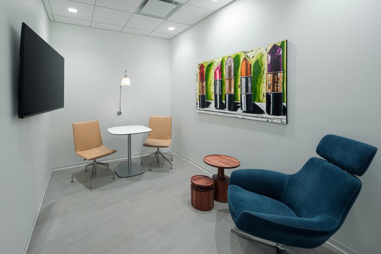 A modern office lunchroom with blue cushioned bench seating and white chairs around small white tables. The room has black cabinets, including a microwave, a sink, and two framed artworks on the wall. A window with blinds lets in natural light.