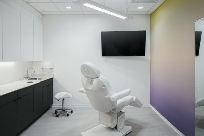 A modern dentist's office with a reclining exam chair facing a wall-mounted TV. The room has sleek white cabinets, a small sink, a cushioned stool, and minimalistic lighting. The wall has a gradient color design.