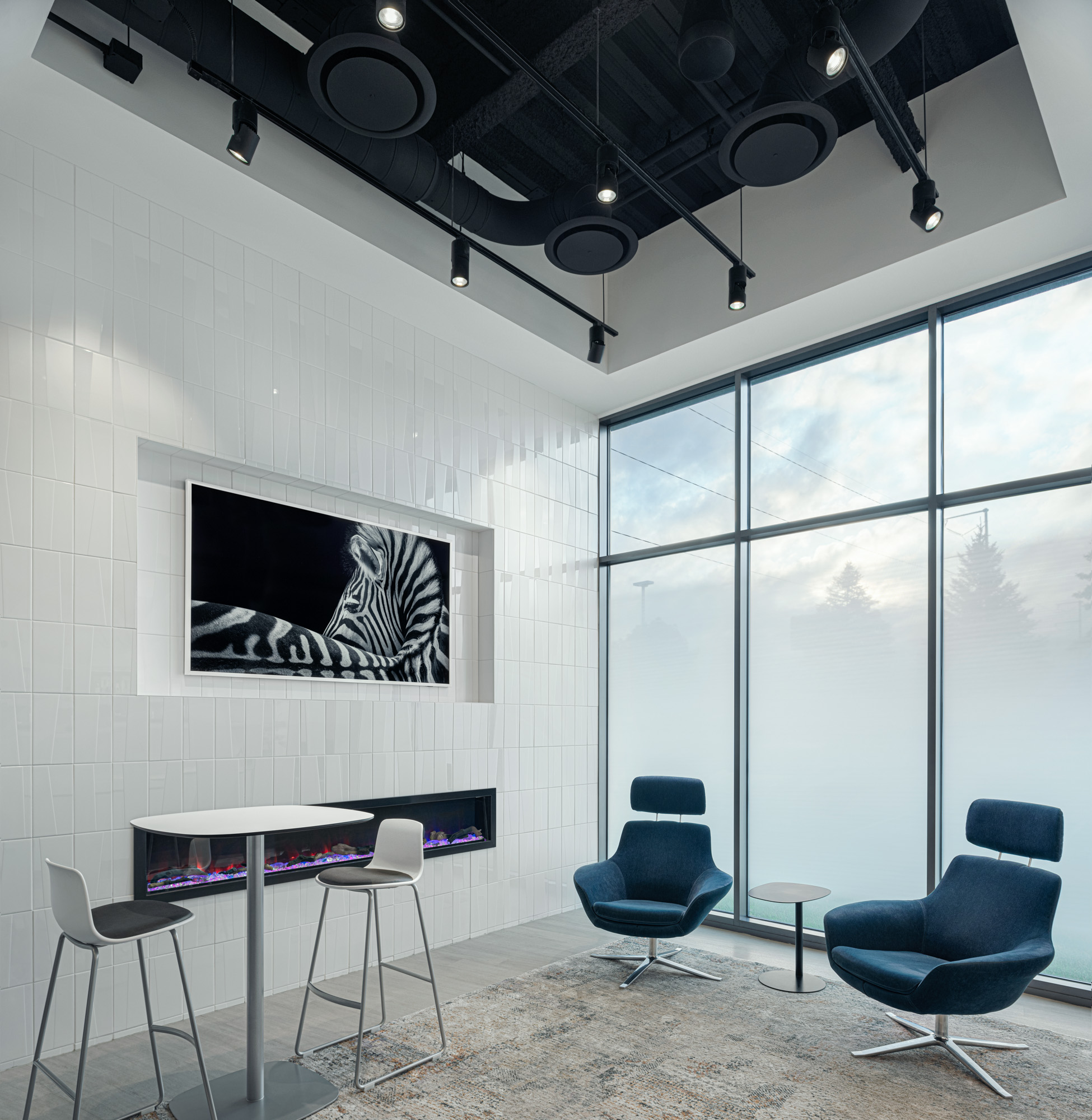 A modern lounge area features two blue armchairs, a rug, a round side table, and a tall table with two barstools. Large floor-to-ceiling windows reveal an outdoor view. A framed zebra portrait and a linear electric fireplace adorn a sleek, tiled wall.