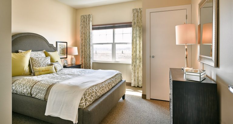 Independent senior living bedroom with large bed, window with light green curtains and cream colored walls