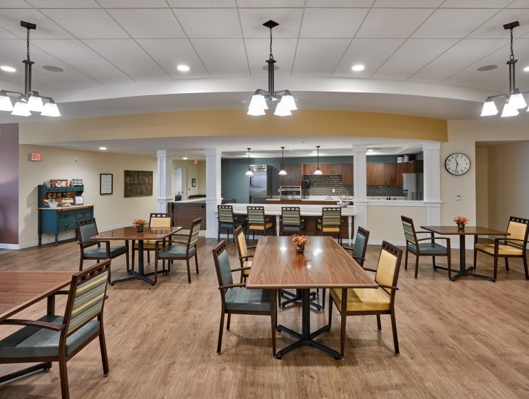 Senior living facility dining area with chandelier lighting, light wood floors and wood top tables with yellow and green accent chairs