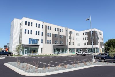 Exterior of building with retail space on the bottom and apartments on the upper 3 levels, an asphalt parking lot and white, cream and gray siding