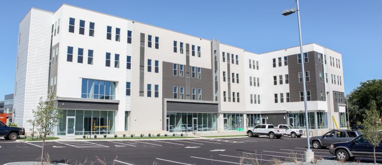 Exterior of building with retail space on the bottom and apartments on the upper 3 levels, an asphalt parking lot and white, cream and gray siding