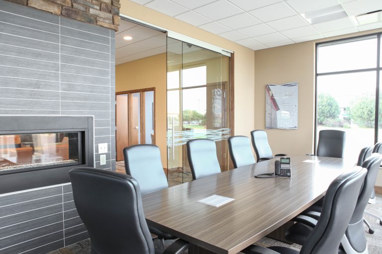 A modern conference room with a long wooden table surrounded by black leather chairs. There's a wall-mounted fireplace on the left, glass doors leading outside, and large windows letting in natural light. A telephone is placed on the table.