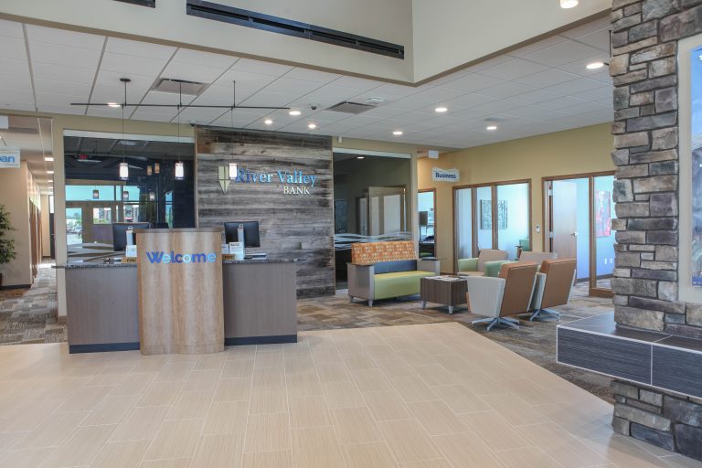 A modern bank lobby with a welcome desk in the center. The space features plush seating, wall accents with stone elements, and clear signage for different areas. Brightly lit, with a mix of carpet and tile flooring.