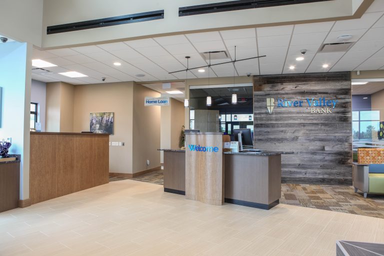 A modern bank lobby with a welcome desk, signage for "Home Loan," and "River Valley Bank" on a wooden accent wall. The interior is bright with neutral tones and contemporary decor.