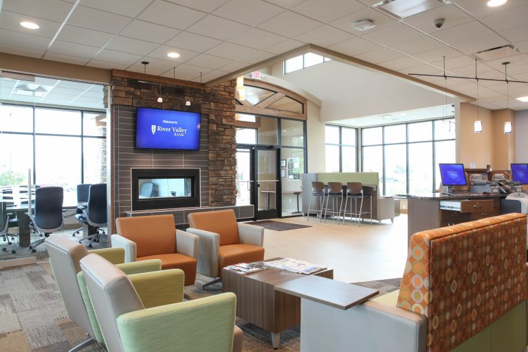 A modern bank lobby with contemporary furniture, including sofas and chairs. A television displaying "River Valley Bank" hangs above a fireplace. Large windows allow natural light, and a reception counter is seen in the background.