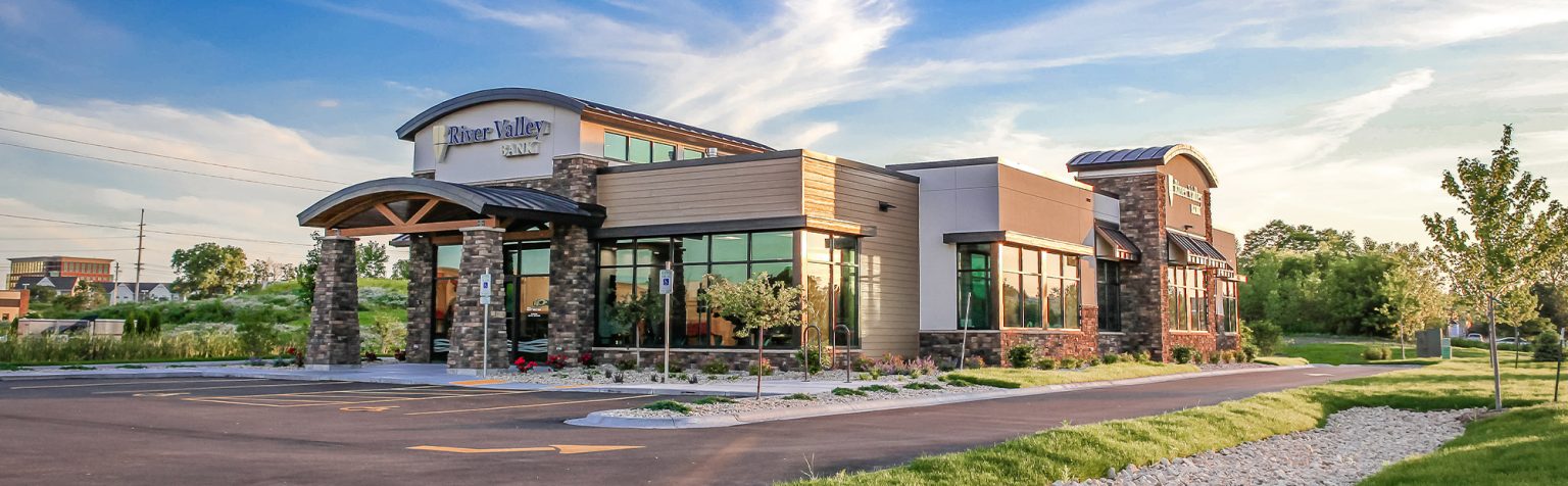 A modern bank with large windows and stone accents under a blue sky with wispy clouds. The foreground features a landscaped area with grass and a paved parking lot.